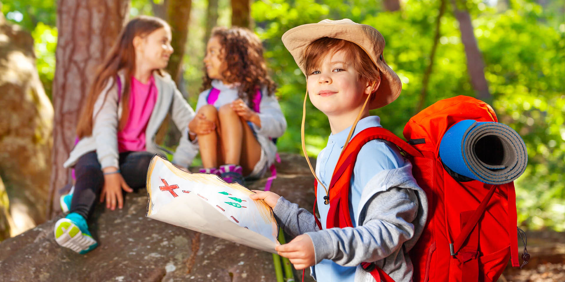 Tipps für die Sommerferien Ein Kind steht in Abenteurer-Ausrüstung und mit einer Schatzkarte im Wald. Hinter ihm sitzen zwei Mädchen auf einem Stein.