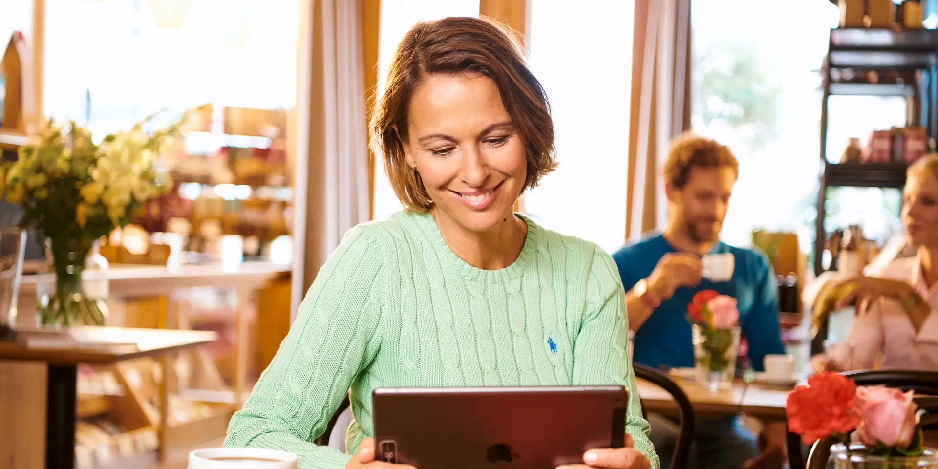 Une femme est assise à une table dans un café, un iPad à la main, en train de lire
