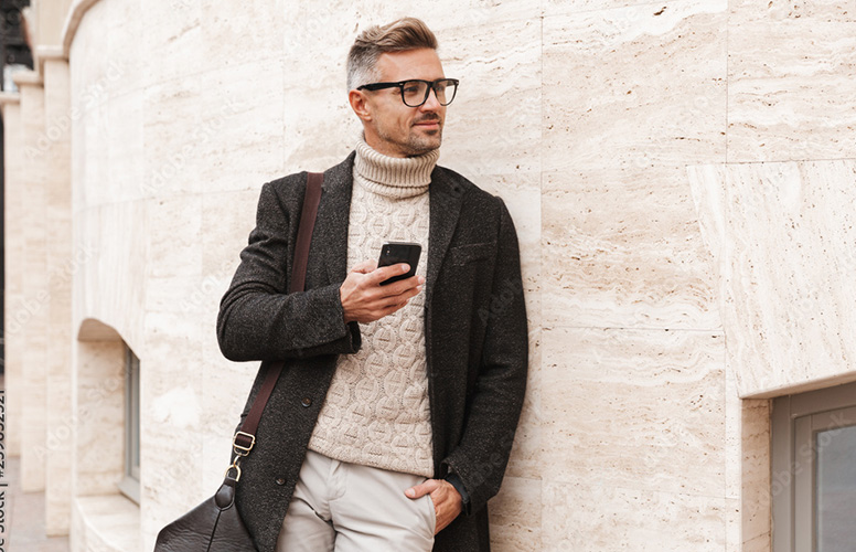 Man in turtleneck sweater leans casually against a house wall with smartphone in hand