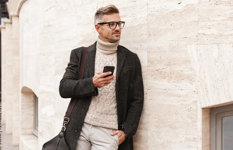 Mann im Rollkragenpullover lehnt lässig an einer Hauswand mit Smartphone in der Hand