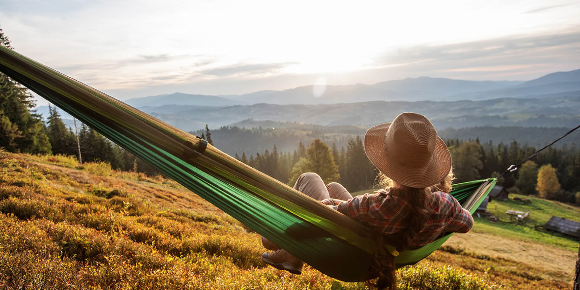 Warum Erholung nach dem Sport so wichtig ist – Frau mit Hit sitzt in einer Hängematte und blickt auf ein Berg-Panorama in der Abendsonne