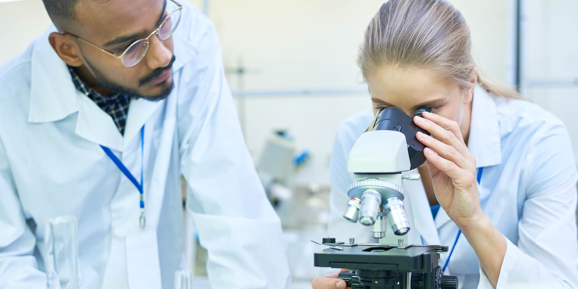Eine Laborarbeiterin schaut durch ein Mikroskop, ein Laborarbeiter steht neben links neben ihr.