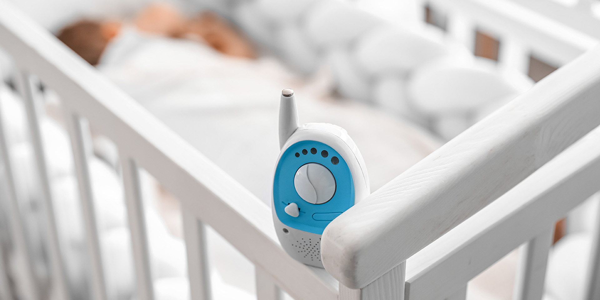 There is a baby monitor on a cot, a baby sleeping in the background