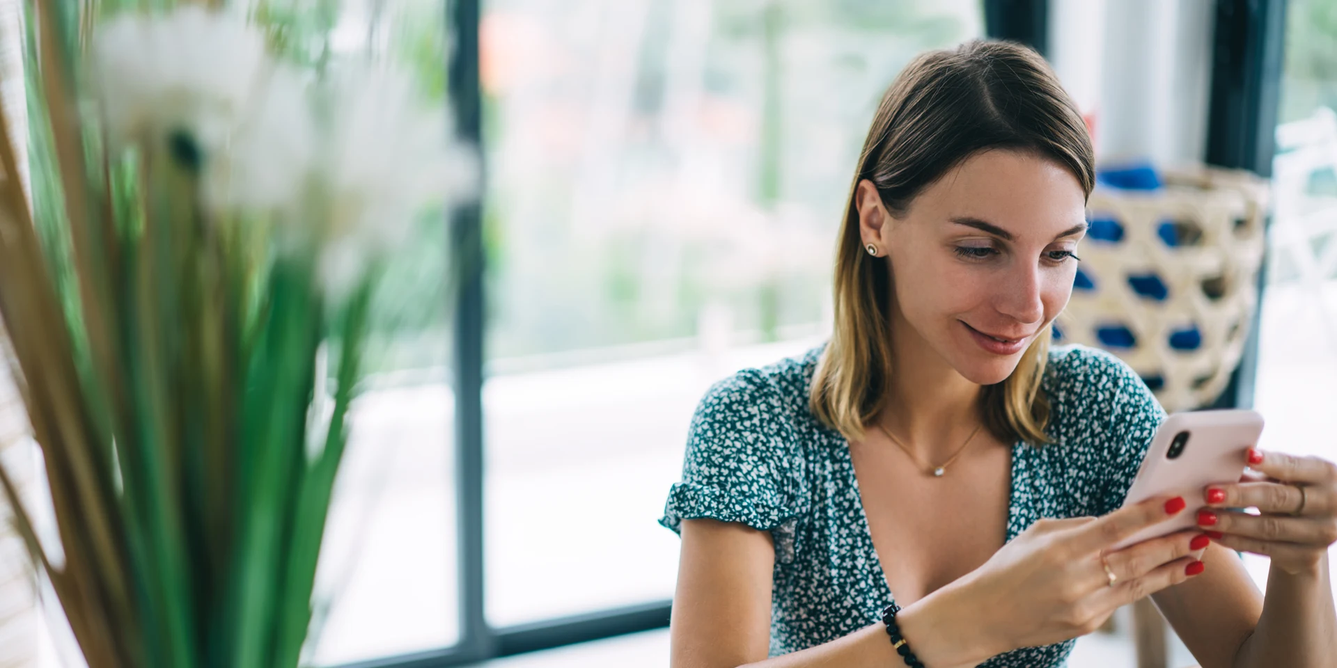 Frau zu Hause schaut auf ihr Smartphone, vor ihr auf dem Tisch steht ein Laptop